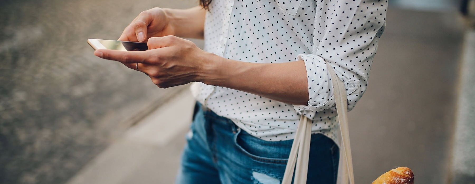 woman walks down the street and texts with a bag of groceries on her arm